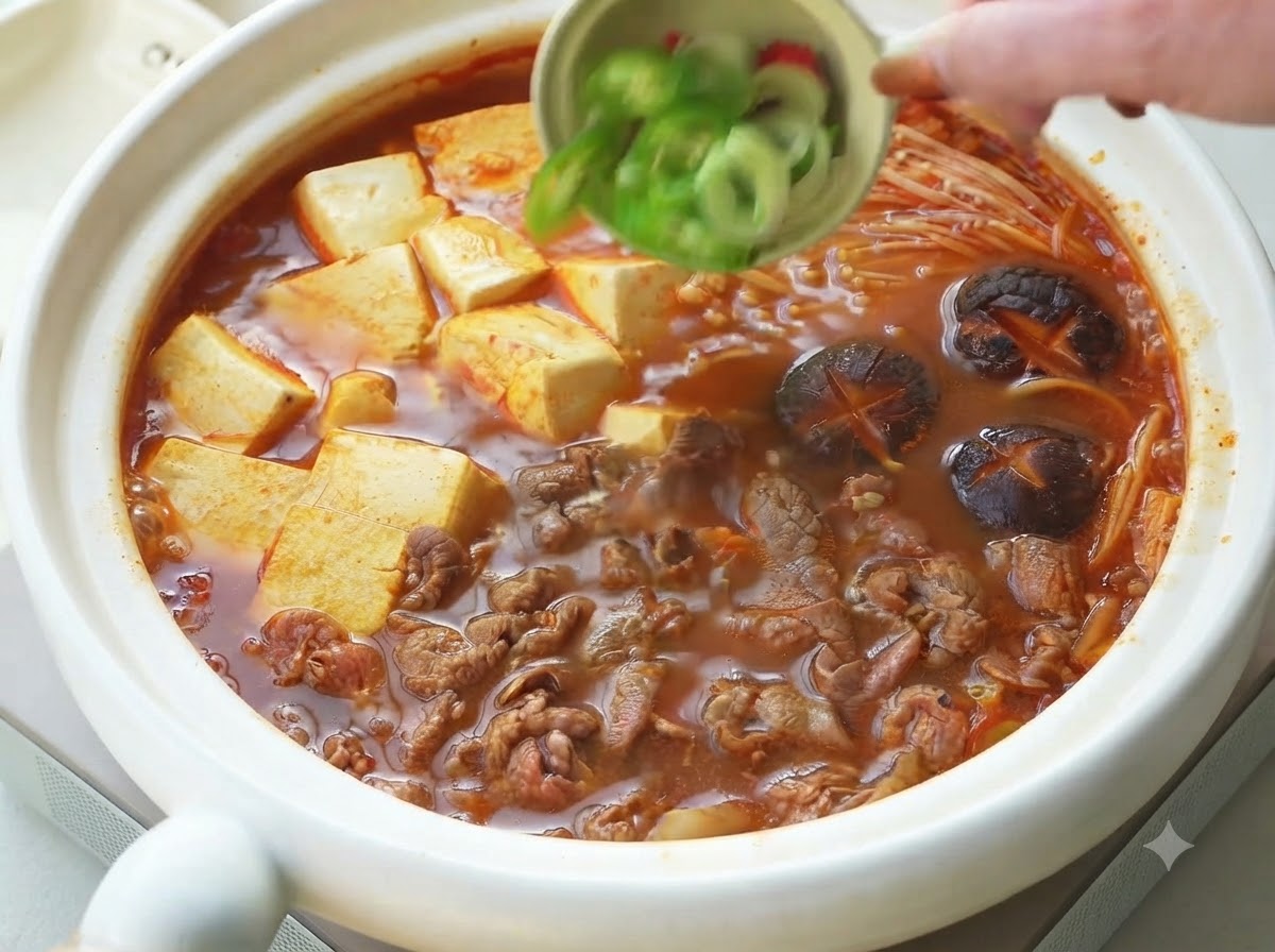 Zielona cebulka i pokrojone papryczki chili wsypywane z małej miski jako dekoracja na ugotowany koreański gulasz wołowy z tofu.