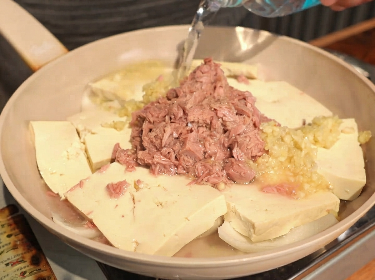 Czysta woda wlewana z butelki na patelnię zawierającą ułożone tofu, cebulę, czosnek i tuńczyka.