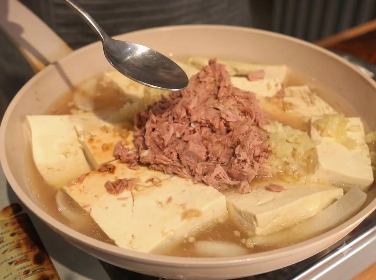 Łyżka trzymana nad patelnią zawierającą tuńczyka, tofu i bulion w trakcie doprawiania.