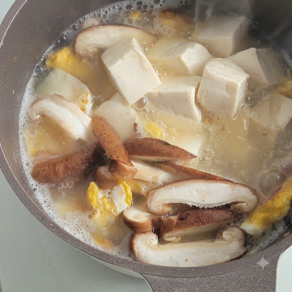 Kostki miękkiego białego tofu gotujące się na wolnym oknie w pikantnym bulionie obok pokrojonych grzybów i kawałków smażonego jajka.