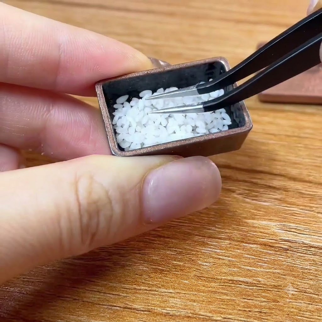 Pęseta umieszczająca malutkie białe ziarna ryżu w miniaturowym brązowym pojemniku.