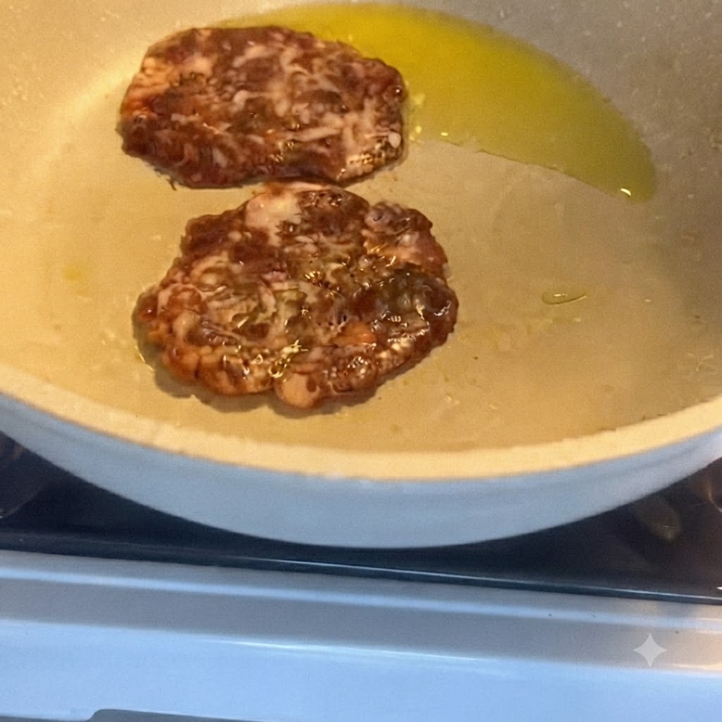 Dwa kotlety wieprzowe smażące się na patelni z olejem, na jednym z nich leży plaster sera.