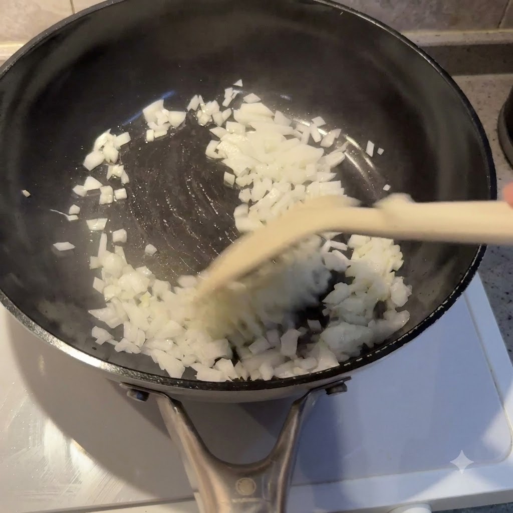 Pokrojoną w kostkę cebulę smażącą się na oleju na czarnej patelni za pomocą łopatki.
