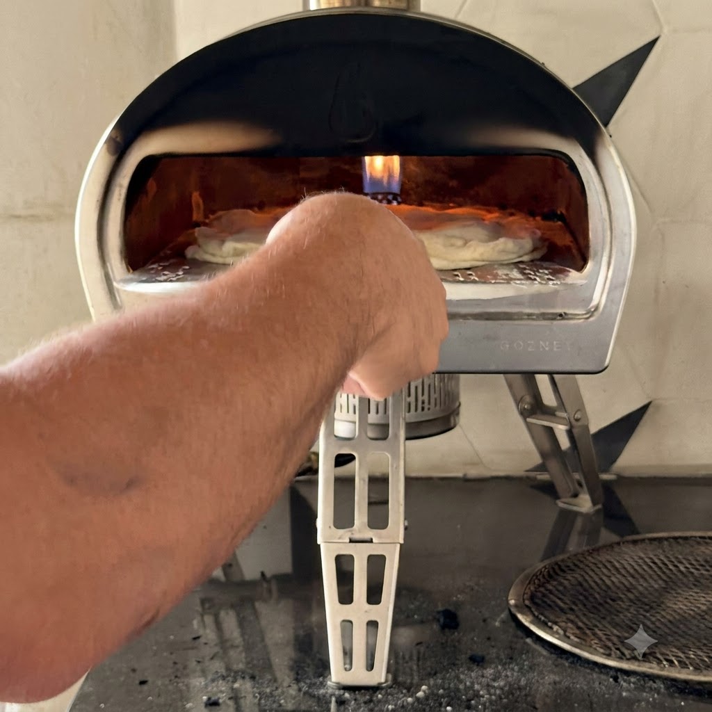 Osoba używająca metalowej łopatki do wsunięcia złożonej surowej pizzy w płonący otwór kompaktowego zewnętrznego pieca do pizzy.